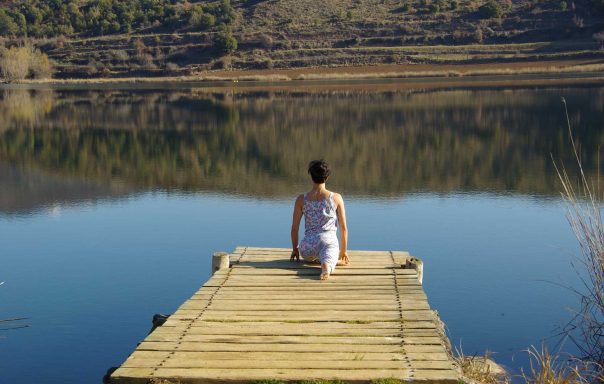 Persona sentada en un muelle de madera frente a un lago tranquilo y montañas reflejadas.