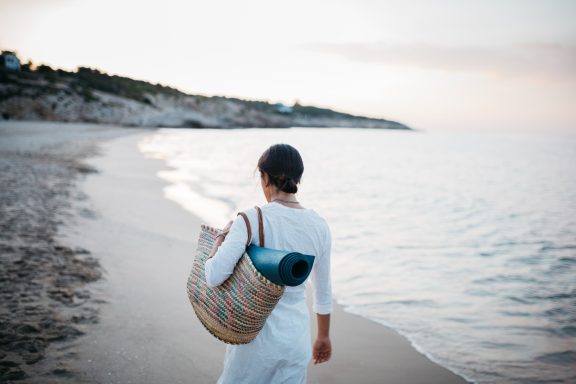 Persona caminando por la playa al atardecer, llevando una mochila y una esterilla de yoga.